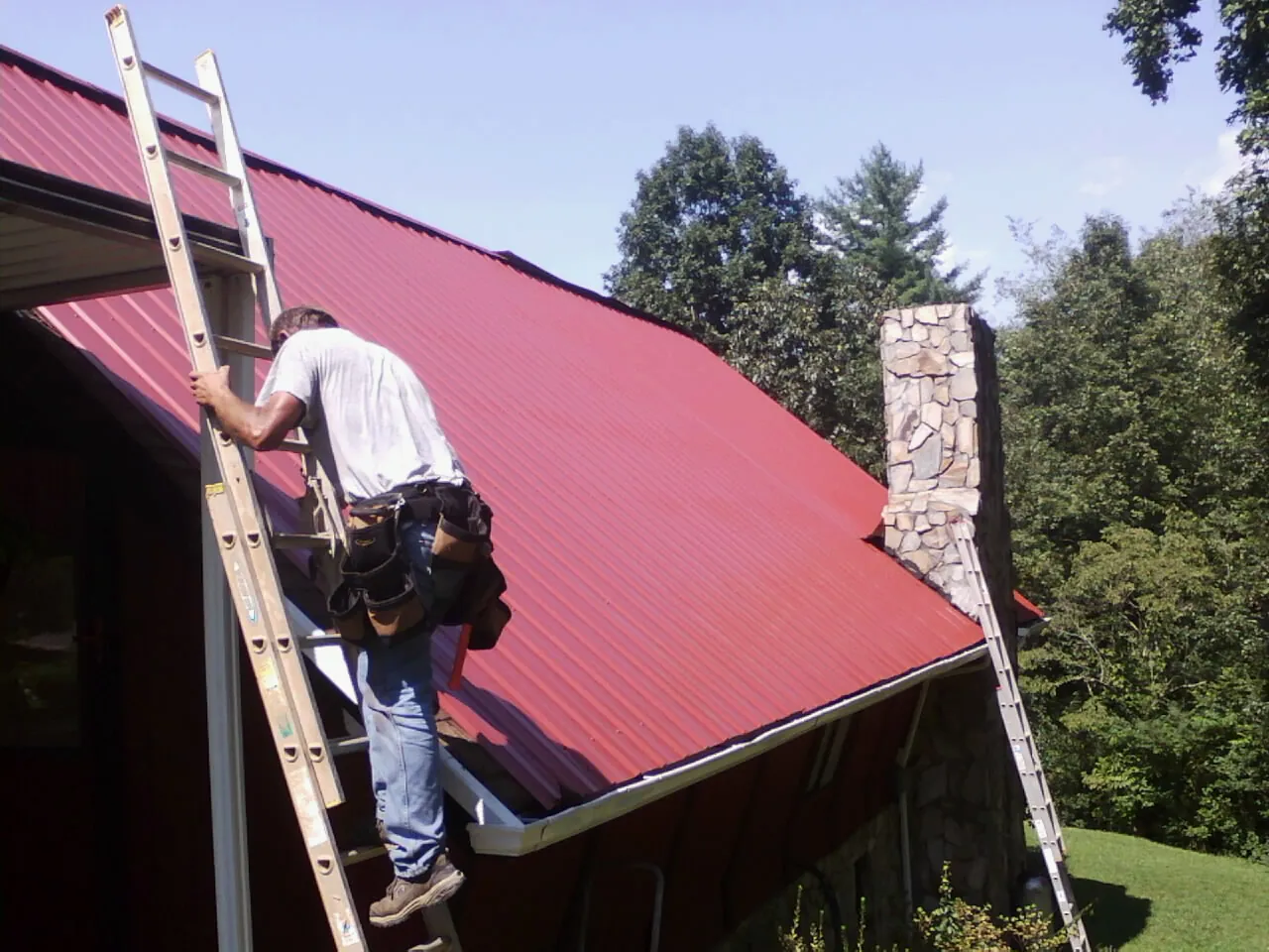 Roofer on a ladder beside a red metal roof and stone chimney — Soffit & Fascia Repair by Roofing Brothers in Pantego
